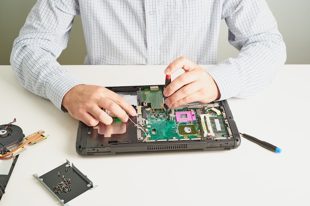 L'homme répare l'ordinateur. un ingénieur de service en chemise répare un ordinateur portable, au bureau blanc contre un mur blanc. Photo Premium