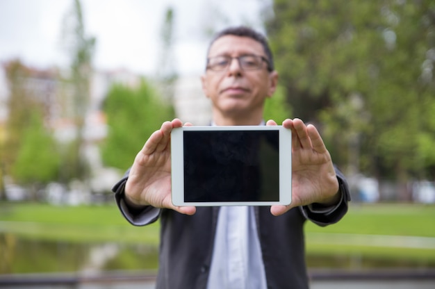 Homme sérieux montrant l'écran de la tablette au spectateur dans le parc Photo gratuit