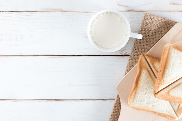Image vue de dessus du pain cuit au four et du pain en tranches avec du lait chaud dans une tasse blanche sur une table en bois blanc, petit déjeuner le matin, frais fait maison, espace de copie Photo Premium