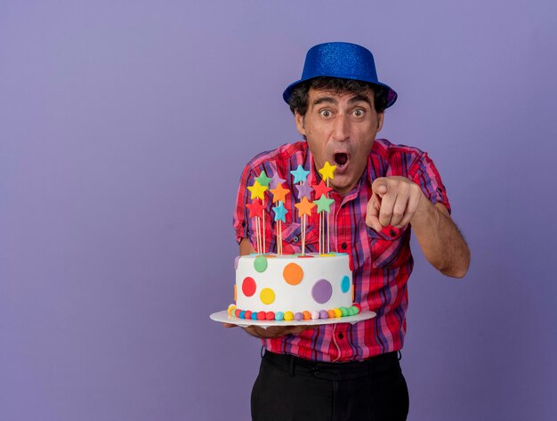 Impressionne Homme De Fete D Age Moyen Portant Chapeau De Fete Tenant Le Gateau D Anniversaire A La Recherche Et Pointant Vers L Avant Isole Sur Mur Violet Avec Espace De Copie Photo Gratuite