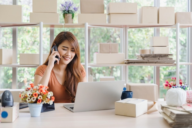Jeune belle femme d'affaires asiatique heureuse en tenue décontractée avec visage souriant appelle pour recevoir les commandes du client avec un ordinateur portable à son bureau à domicile de démarrage, concept pme Photo Premium