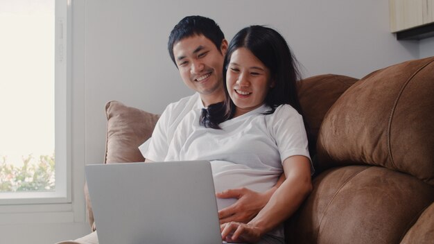 Jeune couple de femmes enceintes asiatiques utilisant un ordinateur portable recherche des informations sur la grossesse. maman et papa se sentent heureux de sourire positif et paisible tout en prenant soin de leur enfant allongé sur un canapé dans le salon à la maison. Photo gratuit