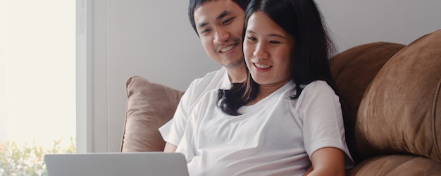 Jeune couple de femmes enceintes asiatiques utilisant un ordinateur portable recherche des informations sur la grossesse. maman et papa se sentent heureux de sourire positif et paisible tout en prenant soin de leur enfant allongé sur un canapé dans le salon à la maison. Photo gratuit