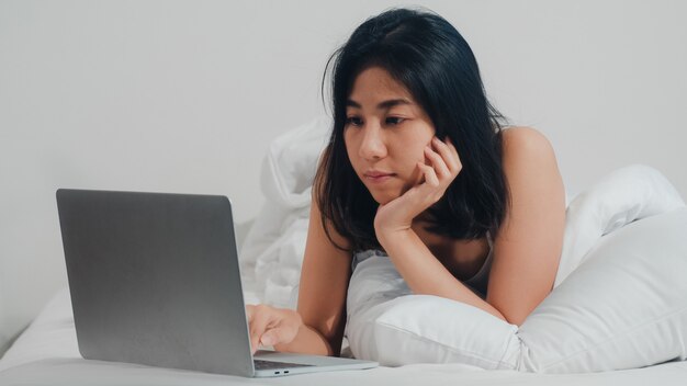 Jeune femme asiatique à l'aide d'un ordinateur portable, vérification des médias sociaux, sentiment heureux de sourire en position couchée sur le lit après le réveil à la maison le matin, jolie fille thaïlandaise souriante se détendre dans la chambre à la maison. Photo gratuit