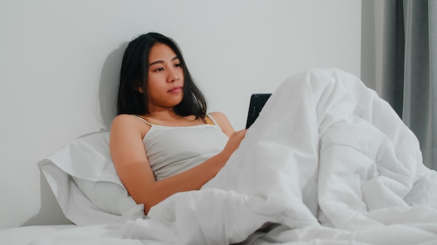 Jeune Femme Asiatique à L'aide De Smartphone, Vérification Des Médias Sociaux, Sentiment Heureux De Sourire En Position Couchée Sur Le Lit Après Le Réveil Du Matin, Belle Demoiselle Hispanique Attrayante Souriante Se Détendre Dans La Chambre à La Maison. Photo gratuit