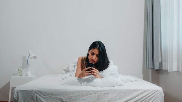 Jeune Femme Asiatique à L'aide De Smartphone, Vérification Des Médias Sociaux, Sentiment Heureux De Sourire En Position Couchée Sur Le Lit Après Le Réveil Du Matin, Belle Demoiselle Hispanique Attrayante Souriante Se Détendre Dans La Chambre à La Maison. Photo gratuit