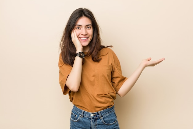 Jeune femme brune contre beige tient sur une paume, gardez la main sur la joue. étonné et ravi. Photo Premium