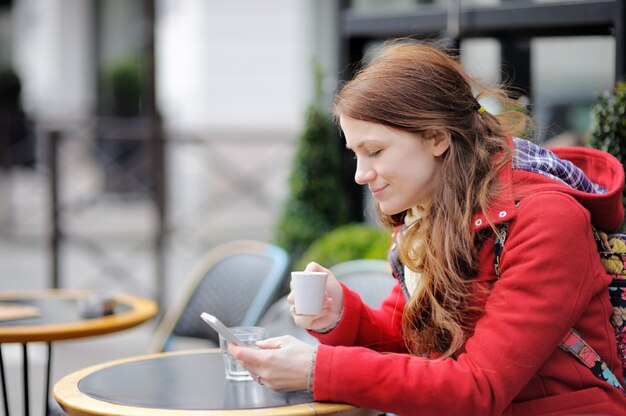 Jeune femme buvant du café et en utilisant son téléphone intelligent dans un café de rue parisien Photo Premium