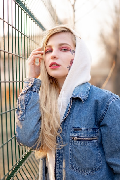 Jeune femme avec des fleurs peintes temporaires sur le visage. adolescente aux longs cheveux blonds porter une veste en jean et capuche Photo Premium