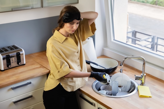 Une jeune femme nettoie dans la cuisine, lave la vaisselle. elle est fatiguée et insatisfaite du fait qu'elle doit le faire. Photo Premium