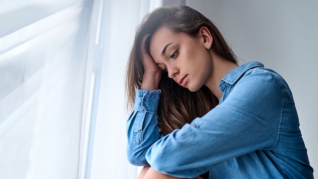 Jeune Femme Pensive Solitaire Triste Dans Une Chemise Tient Sa Tete Avec La Main Est Assis Seul A La Maison Par La Fenetre Pendant La Depression Et Les Soucis Photo Premium