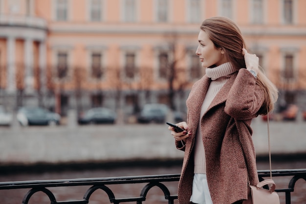 Jeune femme regarde de côté avec une expression réfléchie, tient un téléphone cellulaire moderne, attend l'appel Photo Premium