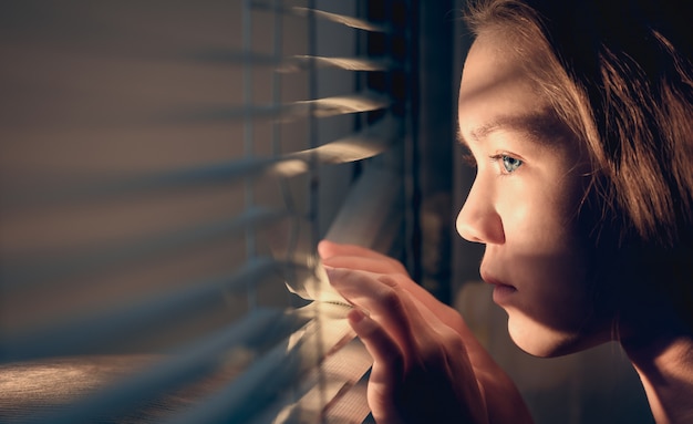 La Jeune Fille Regarde Par La Fenêtre En Poussant Les Stores Avec Ses