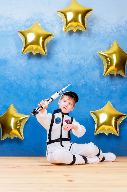 Jeune garçon garçon jouant dans l'astronaute avec une fusée en costume d'astronaute blanc et rêvant de voler dans le cosmos à travers les étoiles en restant à proximité des ballons de l'étoile d'or Photo Premium