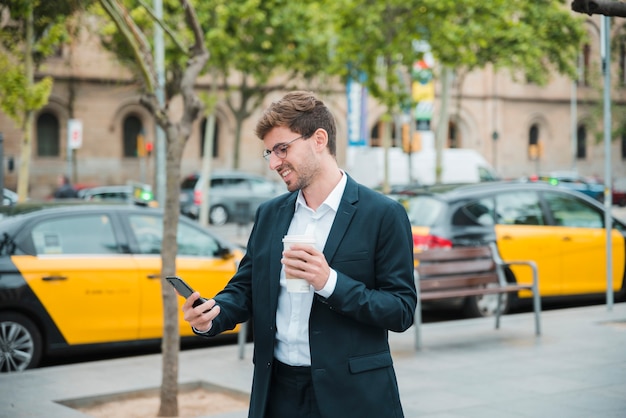 Jeune homme d'affaires tenant une tasse de café en regardant téléphone mobile Photo gratuit