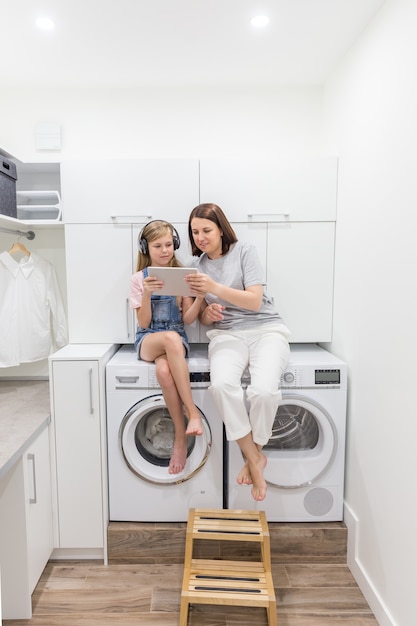 Jeune Mère Et Sa Fille Jouent Avec Une Tablette Dans La Buanderie Assis Sur Une Machine à Laver Photo Premium