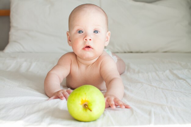 Joli Bebe Joyeux Se Trouve Sur Un Lit Blanc Et Regarde Une Pomme Verte Allongee Devant Lui Photo Premium