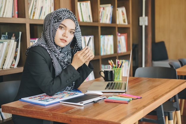 Jolie jeune femme musulmane designer créative à l'aide de tablettes graphiques et ordinateur portable devant l'étagère. Photo Premium