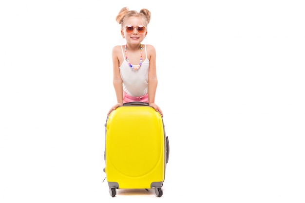Jolie jeune fille en chemise blanche, short rose et lunettes de soleil se tiennent près de la valise jaune Photo Premium