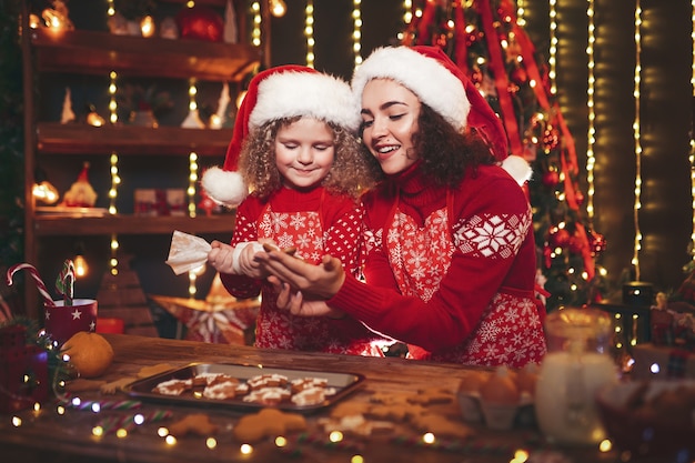 Joyeux noël et joyeuses fêtes. enthousiaste mignonne petite fille frisée et sa sœur aînée à chapeaux de pères noël cuisiner des biscuits de noël Photo Premium