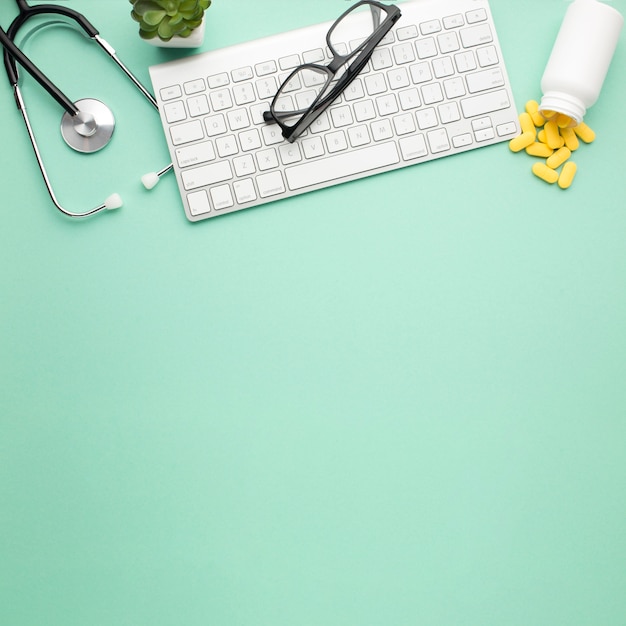 Les lunettes sur le clavier sans fil près des pilules ont renversé la bouteille avant et le stéthoscope sur une surface verte Photo gratuit
