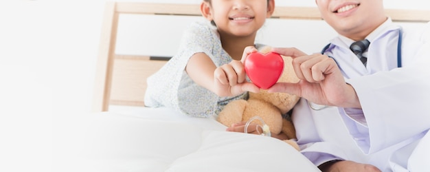 La main de l'enfant asiatique et le médecin de sexe masculin tiennent le coeur rouge allongé sur le lit à l'hôpital. Photo Premium