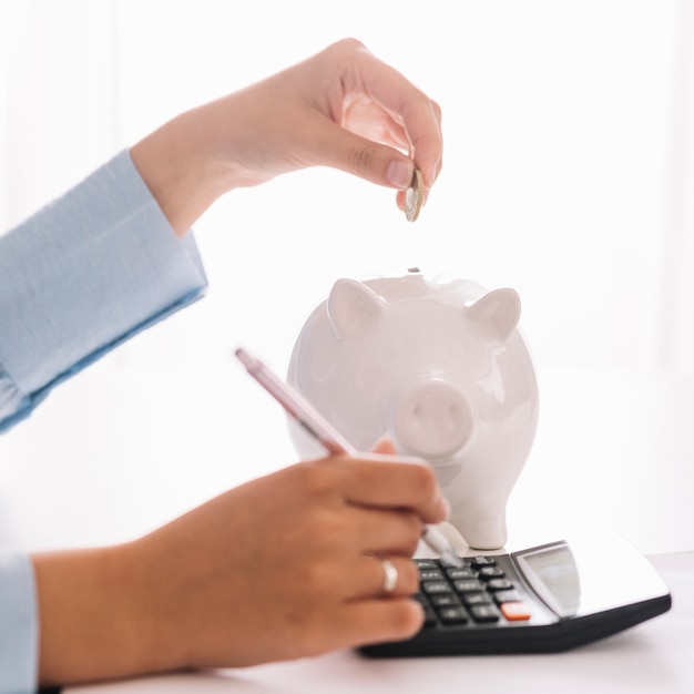 Main de femme à l'aide de la calculatrice lors de l'insertion de la pièce dans la tirelire Photo gratuit