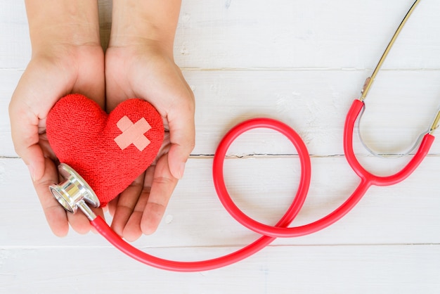 Main de femme tenant un coeur rouge avec stéthoscope sur fond en bois blanc Photo Premium