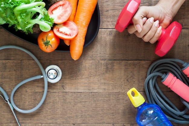 Main de femme tenant un haltère, des aliments sains et des équipements de fitness sur bois Photo Premium