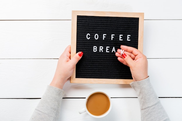Des mains féminines avec des clous rouges tenant un tableau avec des mots pause café près de tasse de café sur une table en bois blanche. vue de dessus Photo Premium