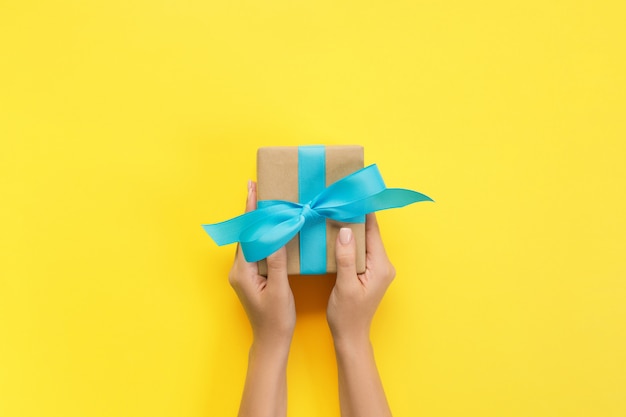 Des mains de femme donnent des cadeaux emballés à la main pour la saint-valentin ou d'autres fêtes en papier avec ruban bleu. boîte à cadeaux, décoration de cadeau sur une table jaune, vue de dessus avec espace de copie Photo Premium
