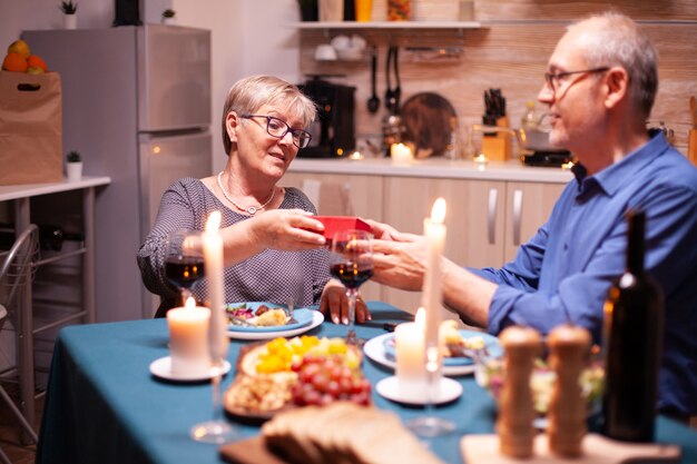 Mari Age Donnant Une Boite Cadeau A Sa Femme Pendant Le Diner Joyeux Couple De Personnes Agees Joyeux Dinant Ensemble A La Maison Savourant Le Repas Celebrant Leur Anniversaire Vacances Surprise Photo