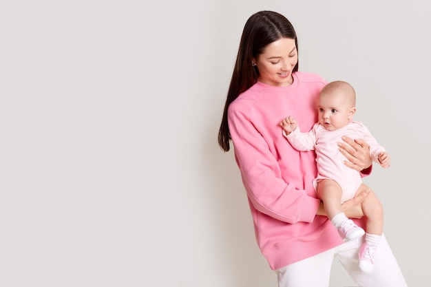 Mere Caucasienne Aux Cheveux Noirs Tenant Bebe Dans Les Mains Et Regardant Sa Fille Belles Robes Feminines Rose Sweat Shirt Avec Bebe Isole Sur Un Mur Blanc Avec Espace De Copie Photo