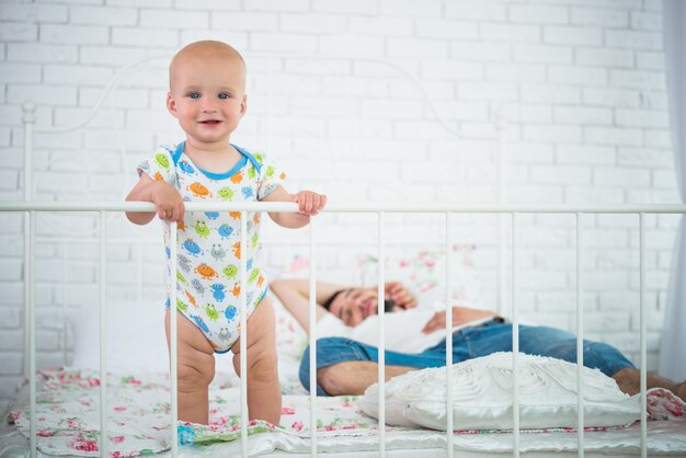 Mignon Petit Bebe Est Debout Sur Le Bord Du Lit Se Tenant A La Tete De Lit Dans Le Contexte Photo Premium