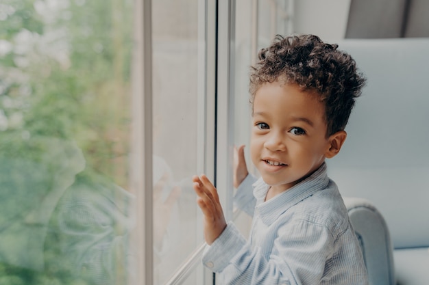 Mignon Petit Garcon Aux Cheveux Boucles Metis En Chemise De Couleur Bleu Clair A Cote D Une Grande Fenetre Photo Premium