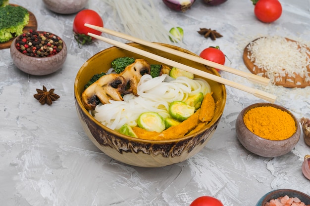 Nouilles de riz cuites; champignon; choux de bruxelles et poulet frit dans un bol avec des baguettes sur une surface texturée en ciment Photo gratuit