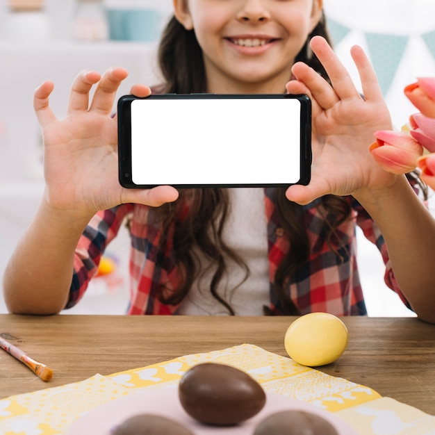 Œufs en chocolat devant une fille montrant un écran blanc sur un téléphone portable Photo gratuit