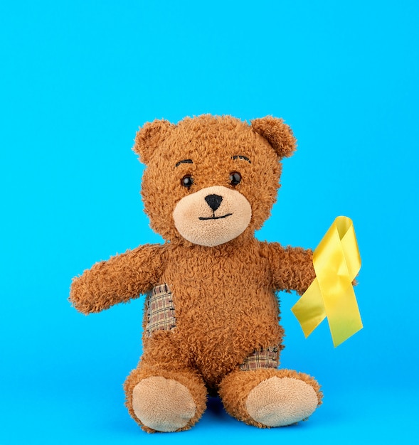 Ours en peluche brun est assis et tient dans sa patte un ruban de soie jaune sur un fond bleu Photo Premium