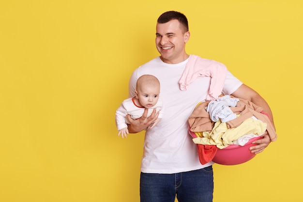 Pere Et Bebe Isoles Sur Un Mur Jaune Beau Regarder Ailleurs Et Rire Tenant Un Enfant Nouveau Ne Et Des Vetements Apres La Lessive Homme Avec Une Expression Heureuse Portant Un T Shirt Blanc