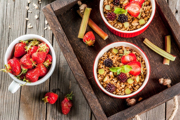 Petit-déjeuner Sain. Crumble D'avoine Granola à La Rhubarbe, Fraises Et Mûres Fraîches, Graines Et Crème Glacée Dans Des Bols Cuits Au Four, Décoré De Menthe, Sur Une Table Rustique En Bois Dans Le Vieux Bac, Vue De Dessus Photo Premium