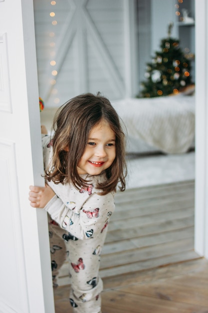 Petite Fille Brune Heureuse En Pyjama Confortable Regarde De La Chambre Des Enfants Le Temps De Noel Photo Premium