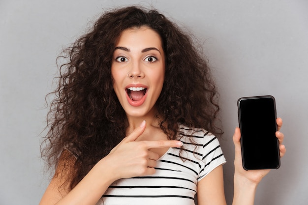 La Photo En Gros Plan De Femme Optimiste Avec Anneau Dans Le Nez, Pointant L'index Comme La Publicité De Son Smartphone, Ce Qui Signifie Un Bon Choix Contre Le Mur Gris Photo gratuit