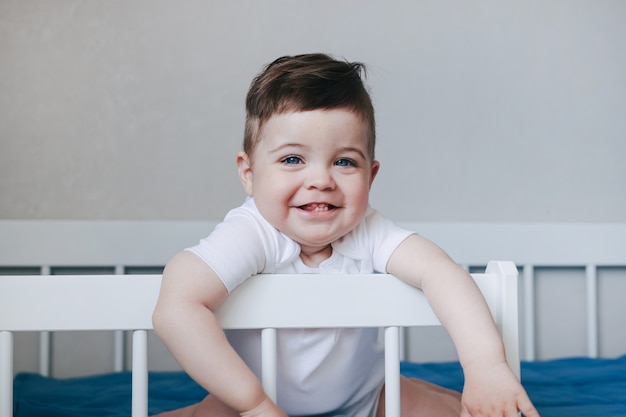 Portrait D Un Bebe Garcon Rampant Et Souriant En Body Blanc Sur Le Lit Dans La Chambre Grands Yeux Bleus Heureuse Enfance Conceptside View Photo Premium