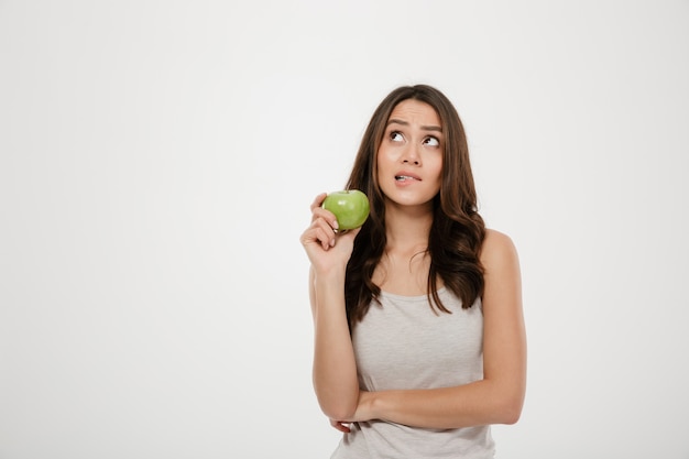 Portrait De Femme Perplexe Regardant Vers Le Haut Tenant Une Pomme Fraîche Verte, Pensant à Des Aliments Sains Isolated Over White Photo gratuit