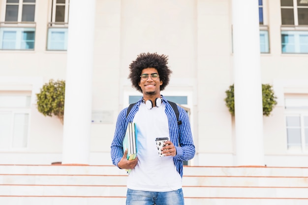 Portrait, heureux, afro, adolescent, étudiant, tenue, livres, et, tasse à café à emporter, debout, devant, collège Photo gratuit