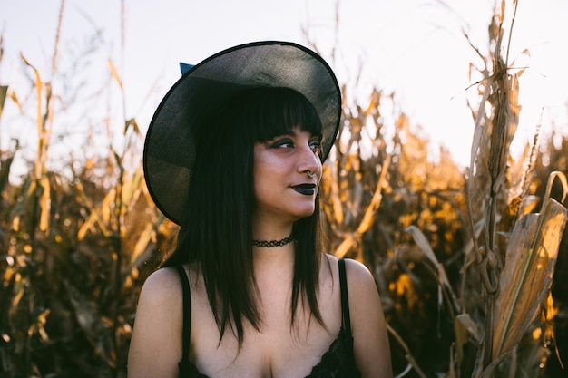 Portrait de jeune fille halloween costume sorcière dans un champ de maïs au coucher du soleil. belle jeune femme sérieuse au chapeau de sorcière aux longs cheveux noirs et aux lèvres noires Photo Premium