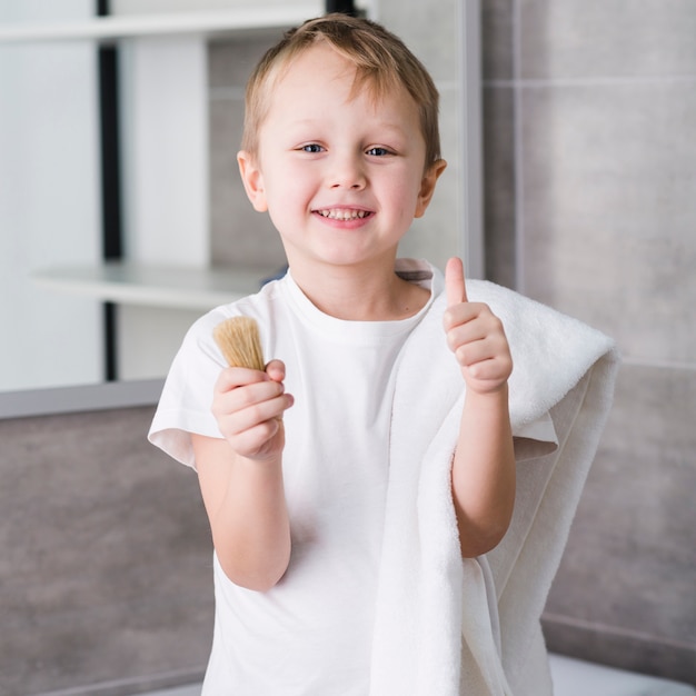 Portrait d'un petit garçon heureux avec une serviette blanche sur son épaule, tenant le blaireau dans la main, montrant le pouce vers le haut de signe Photo gratuit