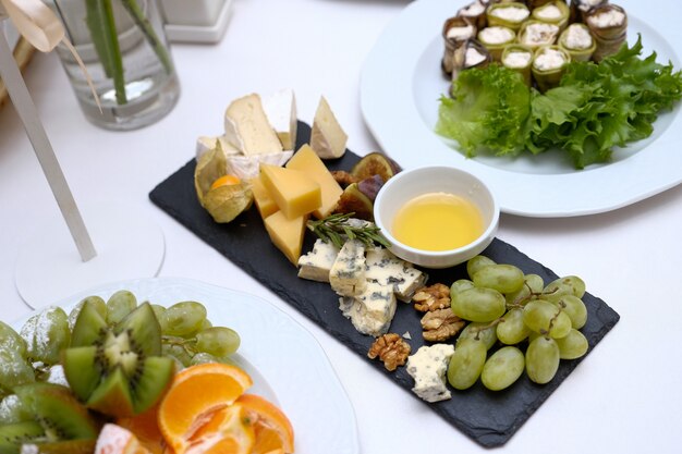 Servir une table de fête avec une variété de plats. morceaux de fromages de différentes variétés, miel dans un bol en verre, fruits, légumes et noix dans un plat en céramique noir sur une table lumineuse. Photo Premium