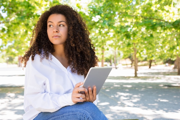 Songeuse jolie jeune femme à l'aide de tablette sur un banc dans le parc Photo gratuit