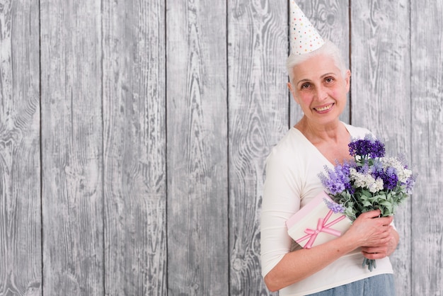 Souriant Anniversaire Femme Tenant Une Boite De Cadeau Et Bouquet De Fleurs Pourpres Devant Fond En Bois Gris Photo Gratuite